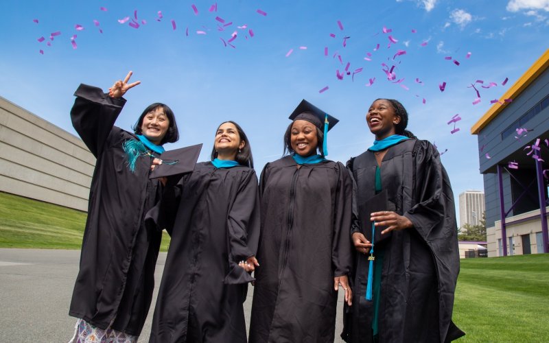 Four graduates in black gowns and caps celebrate outdoors under a blue sky. Purple confetti falls as they smile, exuding joy and accomplishment.