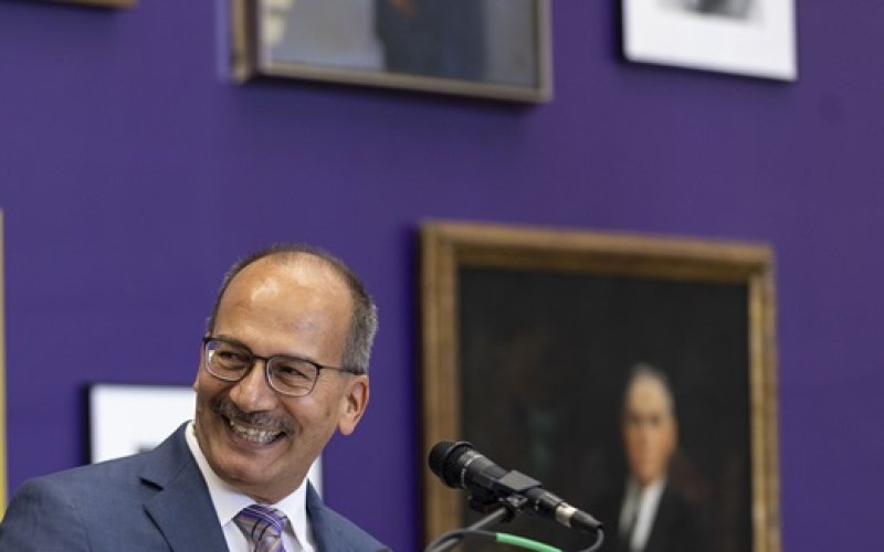 A smiling man in a blue suit stands before a microphone at a lectern in front a purple wall with framed portraits on it.