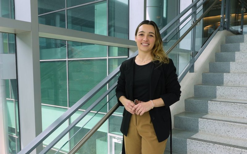 A young woman with brown-blond ombre hair, wearing a black top, black blazer and khaki slacks, smiles for a portrait in a stairwell with glass walls and gentle natural light.