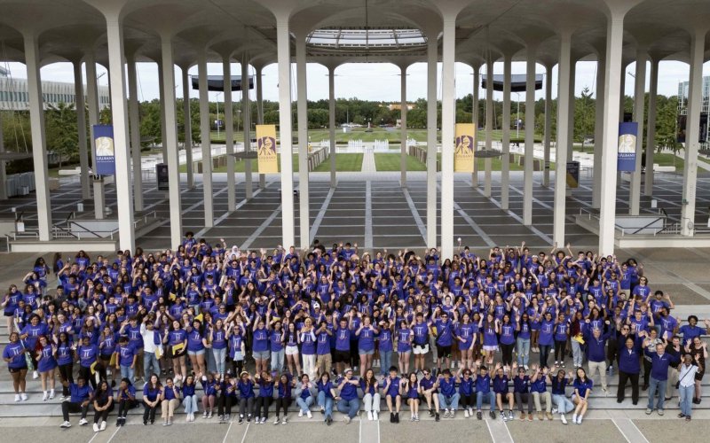 A few hundred college students in purple T-shirts wave while posing for a group photo in front to a pillared plaza with a fountain in the background
