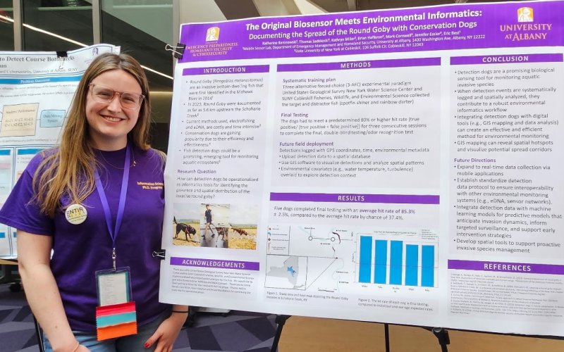 A woman stands next to a research poster titled "The Original Biosensor Meets Environmental Informatics" at a science event, presenting findings on round goby detection with conservation dogs.