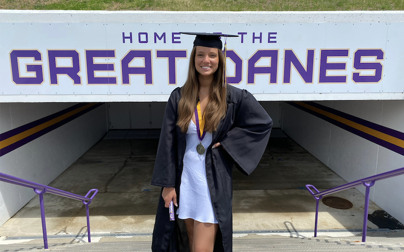 A young woman with long brown hair in a white dress and black graduation cap and gown poses in front of a sign that says "Home of the Great Danes"