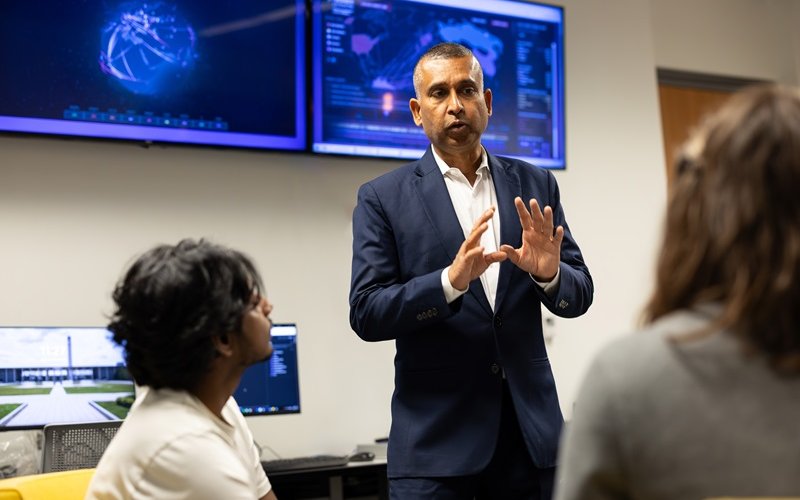 A man in a blue suit and white shirt stands in front of computer screens depicting world maps gesturing with his hands in front of his body while talking to two students.