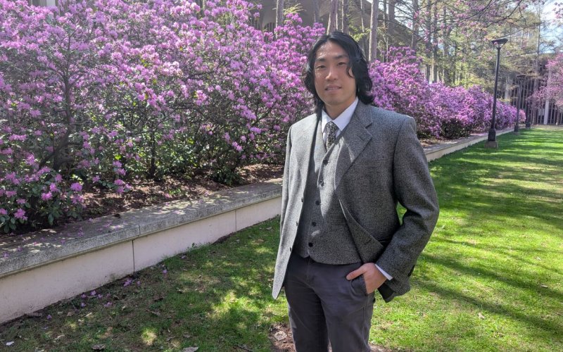 A young man with dark hair wearing a gray blazer and gray khaki pants poses for a portrait in front of a magnificent hedge of purple azaleas in full bloom on a sunny day.