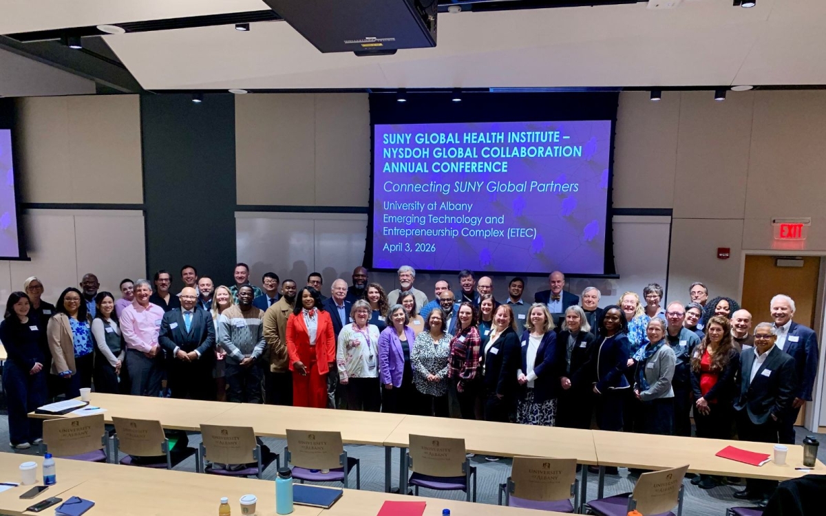 Conference members stand together in a conference room, in front of a presentation screen.