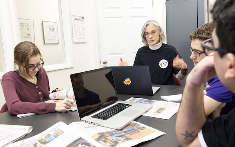 A group of students sit at a table with laptops discussing journalism and articles with a professor wearing a blue sweater.