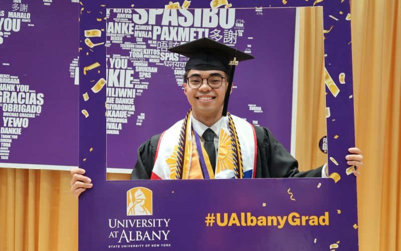 A UAlbany student poses in a purple picture frame, wearing academic regalia.