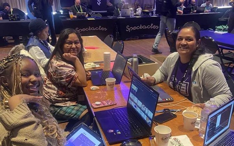 Three smiling women sit with laptops at a table strewn with cellphones, paper coffee cups and water bottles.
