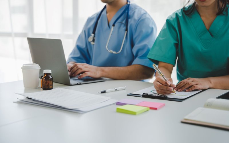 Two people wearing scrubs sit at a white desk. One is working on a laptop, wearing a stethoscope. The other is writing notes on a clipboard. Papers, post-it pads, and several medicine bottles are also on the table.
