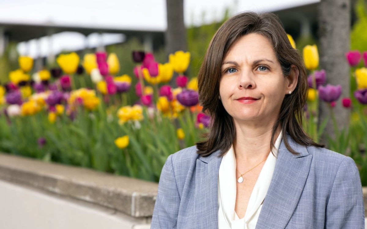 A woman with brown hair wearing a blue-gray blazer and white blouse poses for a portrait in front of a flower bed full of yellow, purple, and magenta tulips in full bloom.