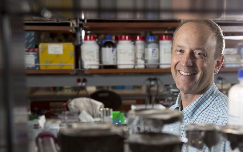 A man smiles for a portrait taken between two shelves crowded with biology lab equipment.