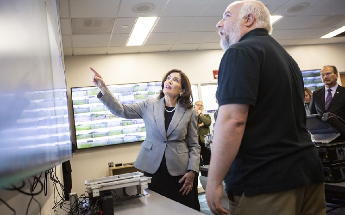 A woman with brown, shoulder-length brown hair and wearing a gray blazer points to a wall-mounted computer screen while a man in a black polo shirt looks on from the foreground.