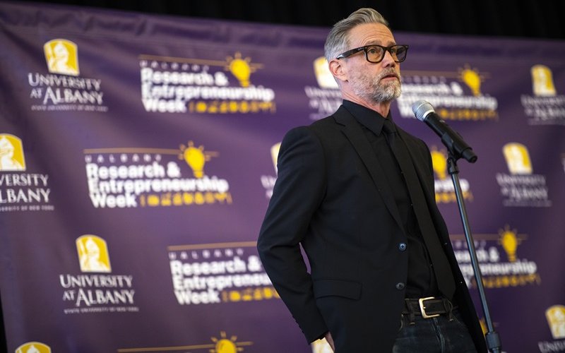 A bearded man with gray/black hair wearing dark-rimmed glasses and wearing all black standing at a microphone in front of a purple backdrop with the words "University at Albany" and "Research and Entrepreneurship Week" on it.