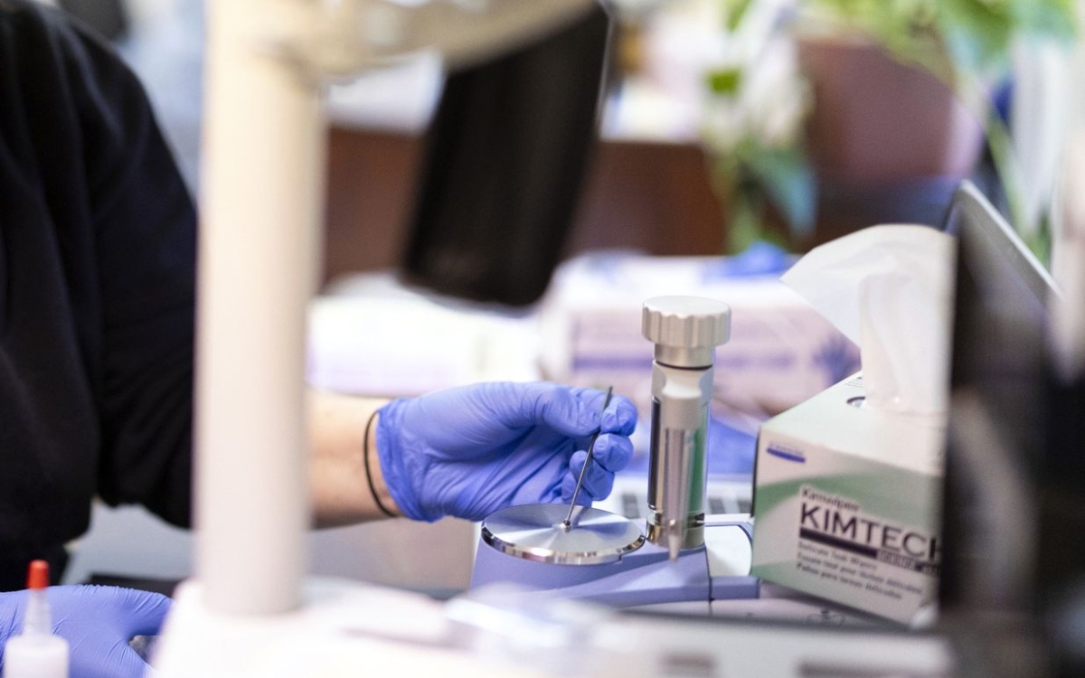 Using a small metal instrument, a person wearing purple latex gloves arranges a tiny amount of a white substance in the center of a round silver platform attached to a piece of lab equipment.
