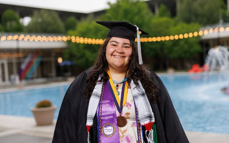 A young woman wearing graduation garb smiles for a portrait in front of Albany’s main fountain. Her cap carries a gray tassel and she is wearing multiple colorful sashes and cords as well as a medal. One of the Sashes us purple satin and says “Middle Earth Peer Assistance Program” with the Middle Earth logo. 
