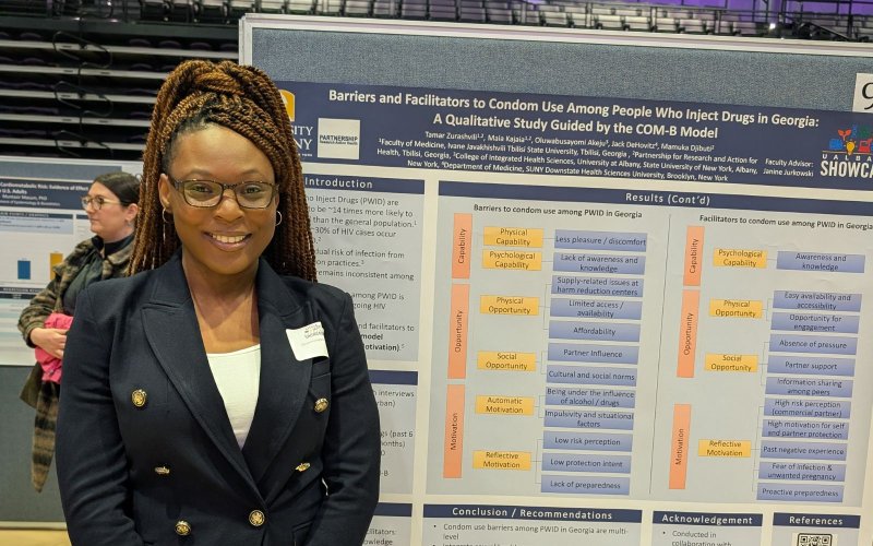 A portrait of Busayo at UAlbany Showcase. She stands in front of her academic poster, which displays information about her work in Georgia.