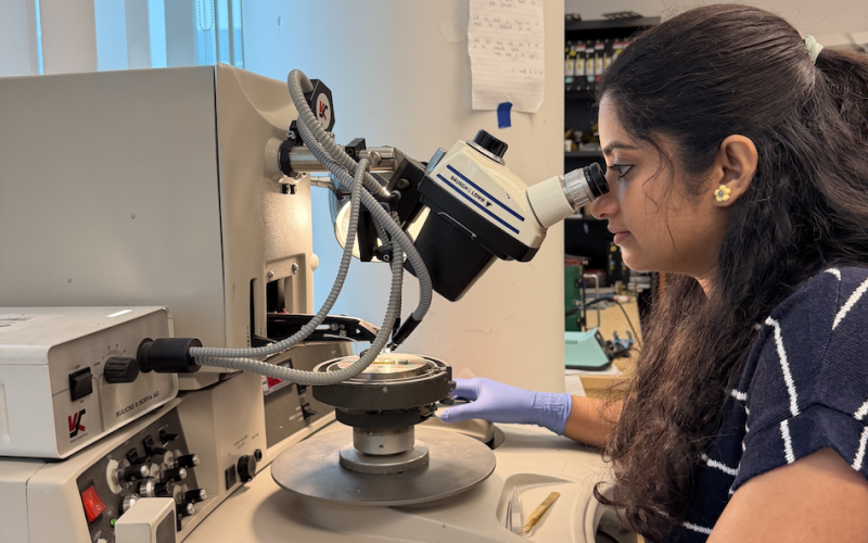A woman with long dark hair in a blue and white striped blouse looks into a microscope in a laboratory