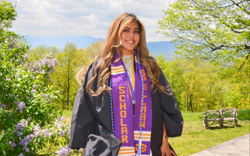 A young woman with long gold-brown hair smiles for a portrait wearing a black graduation gown. Around her neck, she is wearing a blue and white cord, a medal hung on a purple and gold ribbon, and a purple sash that says “Spellman Scholar.” She is standing outside with a blooming lilac bush and greening springs tree in the background. The Catskill mountains are visible off in the distance.