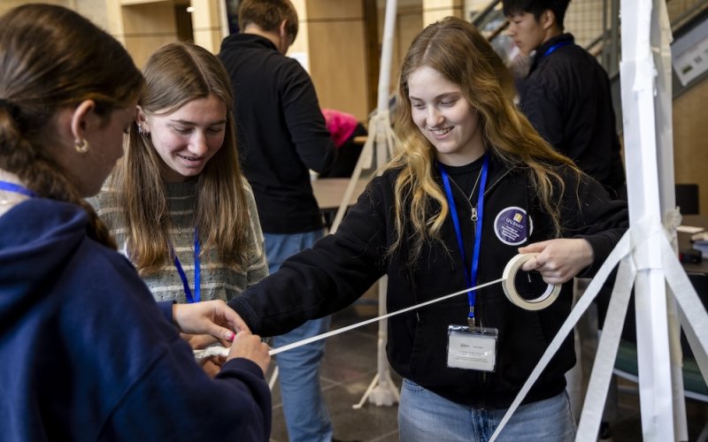Three smiling teen girls hold onto a spool of masking tape as they attempt to build a freestanding paper tower.