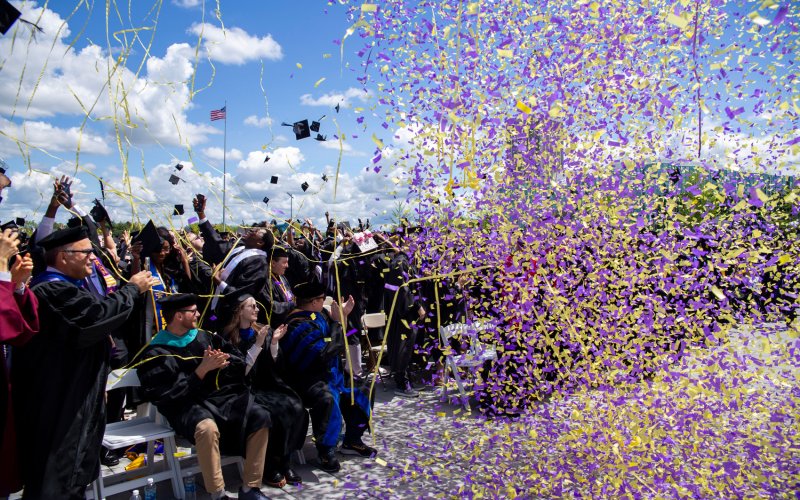 Purple and gold confetti and streamers fall from the sky as graduates clap and smile at UAlbany's Commencement.