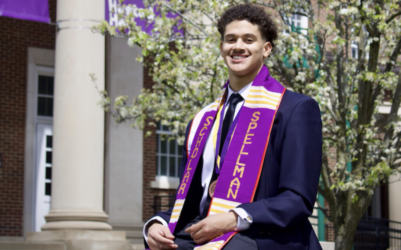 A young man with short curly hair in a suit and purple sash that reads "Spellman Scholar" sits outside on a sunny spring day.