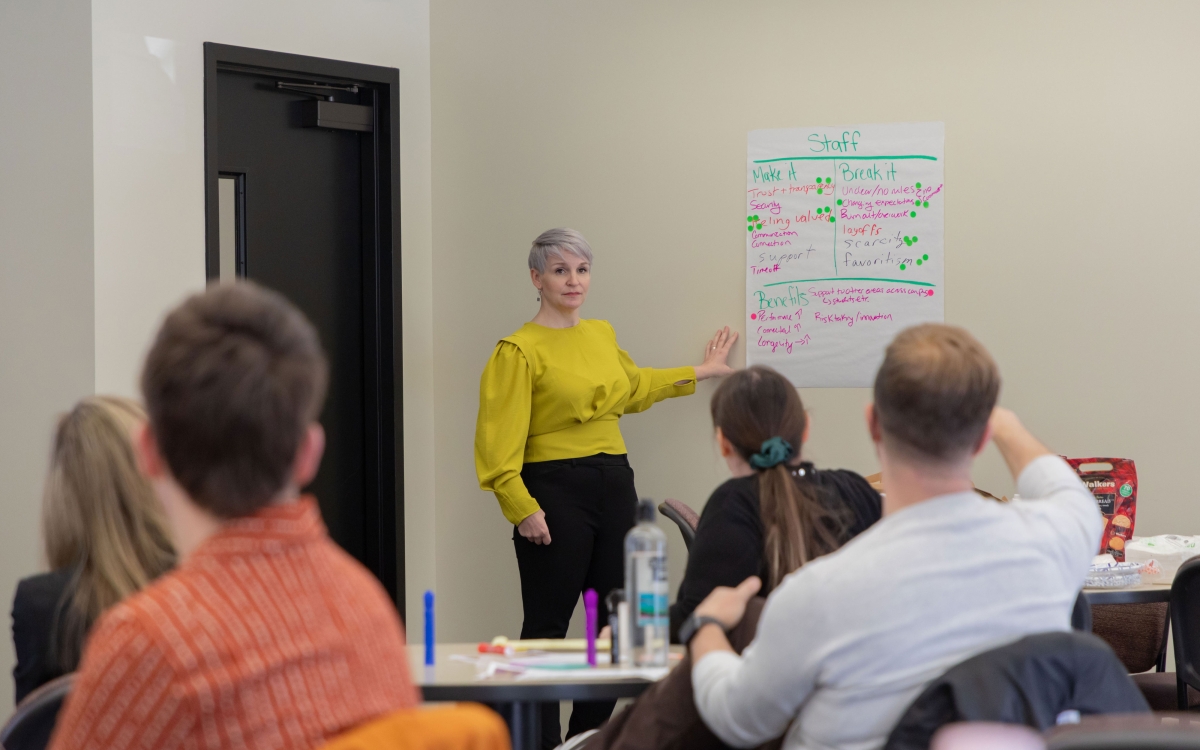 An instructor speaks to a classroom full of students, pointing to a large piece of paper with information on the wall.