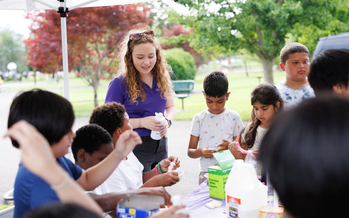A woman in a purple shirt stands in a group of children making slime in a park under a pop-up tent