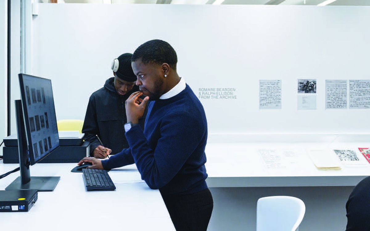 Two students view an archive of materials in a white study space with a sign that reads Romare Bearden & Ralph Ellison: From the Archive on the wall behind them.