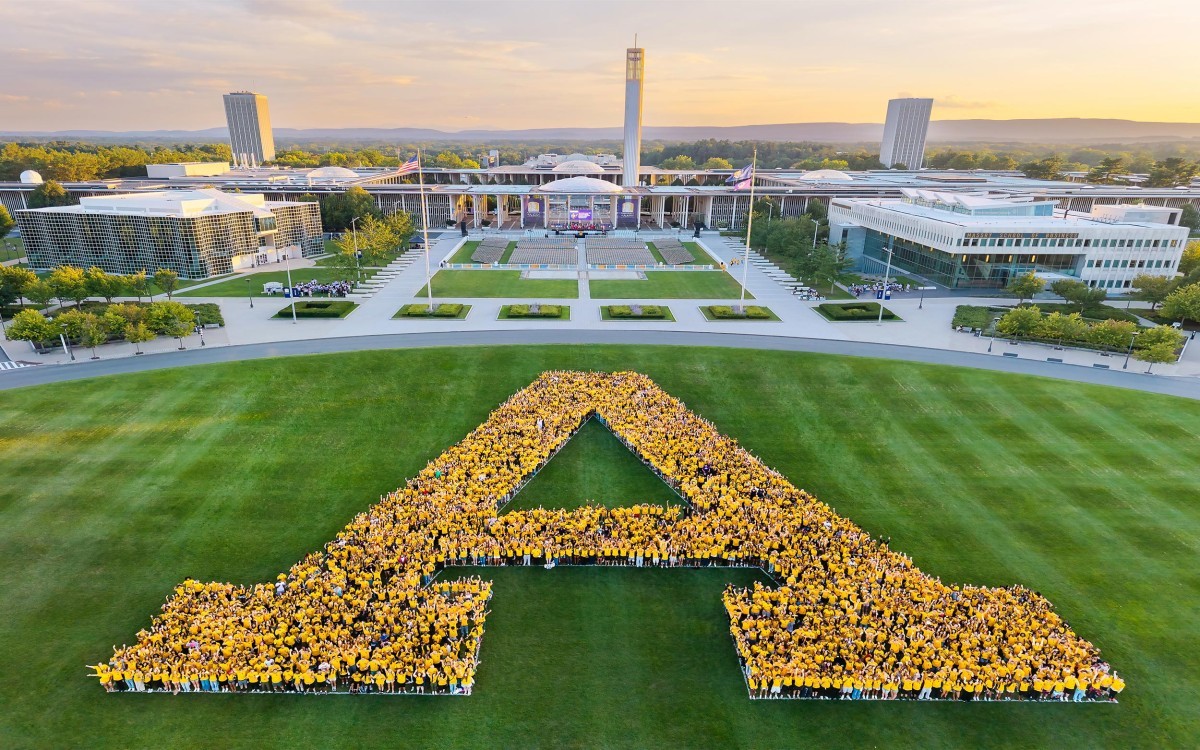 A large group of students wearing yellow t-shirts gather in a huge A on a grass field with the UAlbany campus in the background