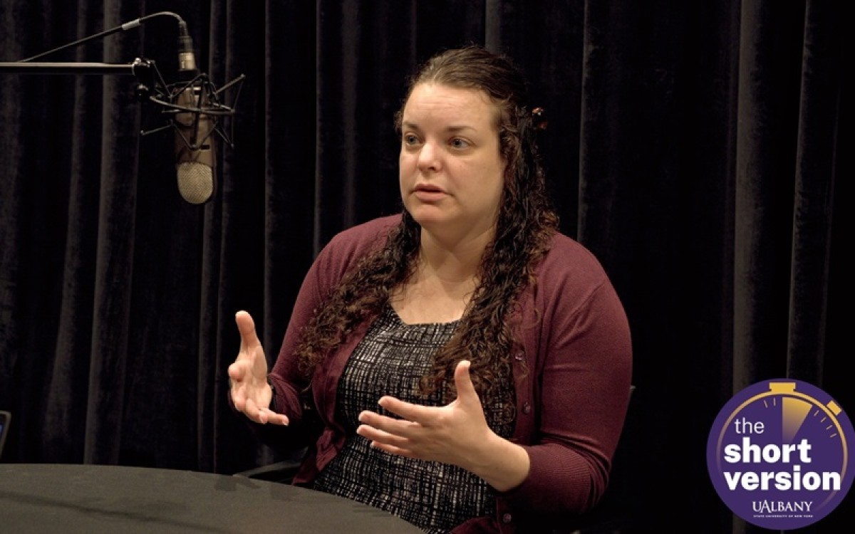 A woman in a maroon sweater sits in front of a microphone against a black background while she gestures with her hand. The words "the short version" are in a purple circle in the lower righthand corner.