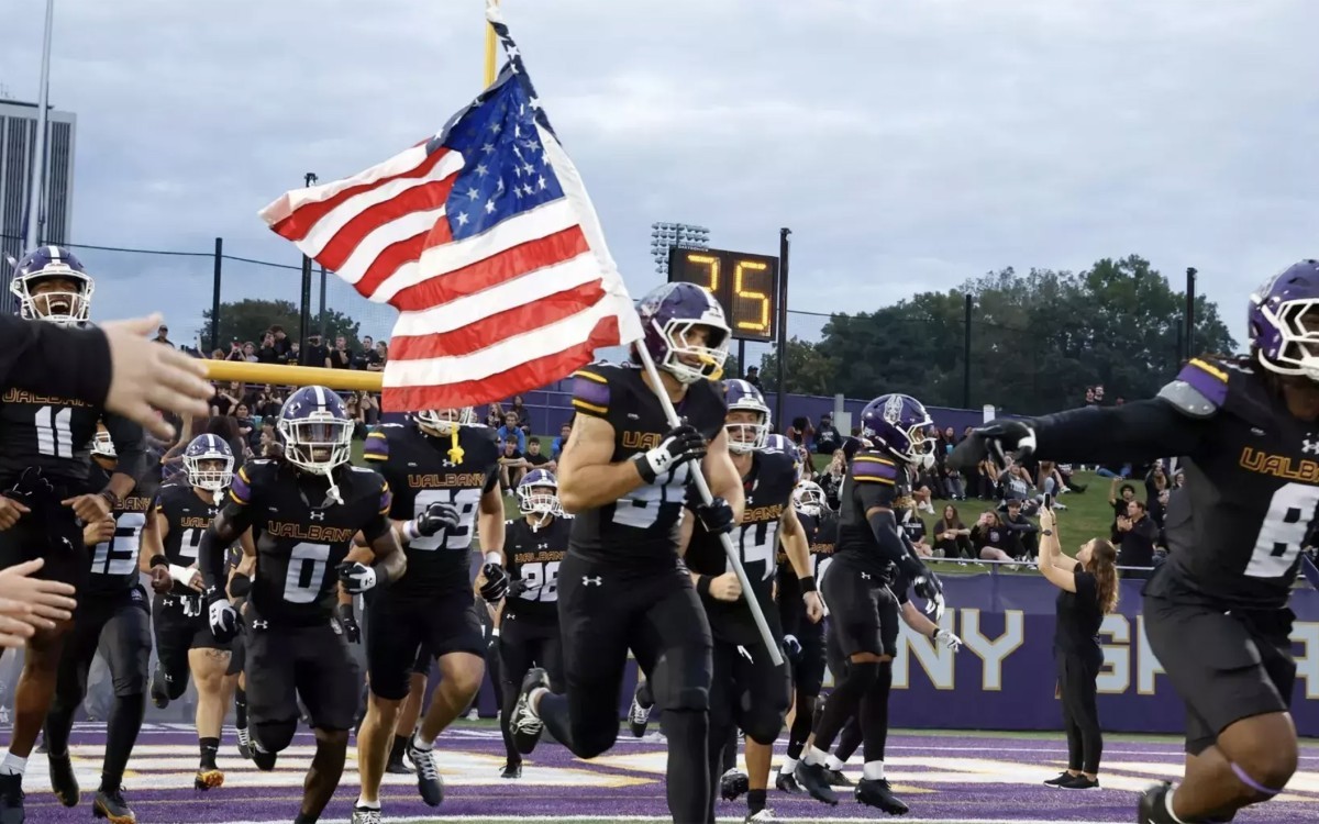 A football player in a black uniform with the word UALBANY on the front carries the American Flag onto the field surrounded by teammates.