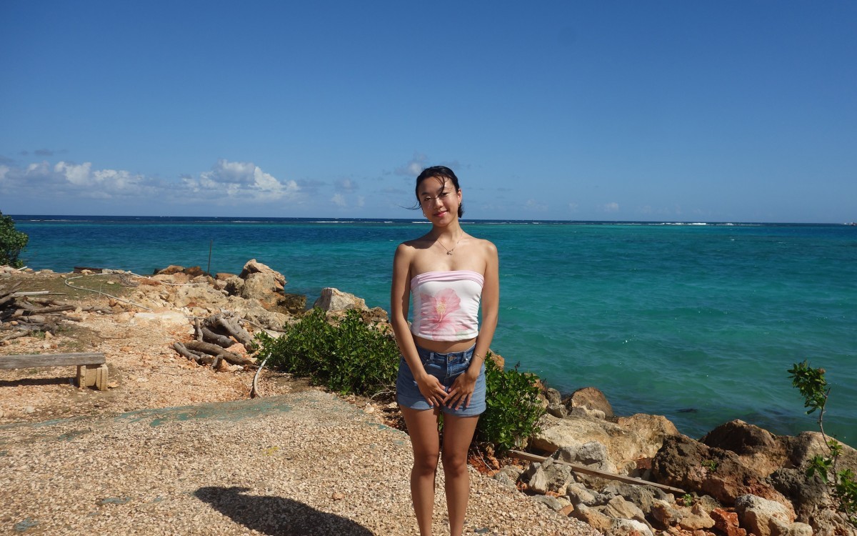 A young student stands along the shore of an Ocean bay as deep blue waters come up to the rocky shoreline behind her in Discovery Bay, Jamaica.