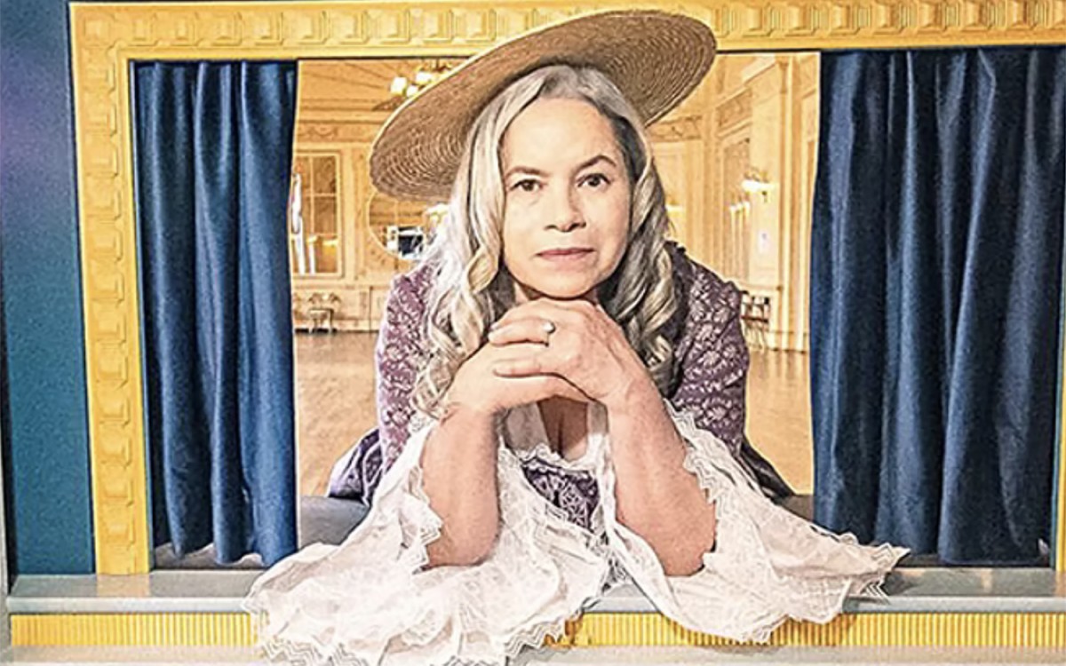 A woman  with gray hair wearing a lace and flower dress and a hat  leans over a railingin front of a curtain.