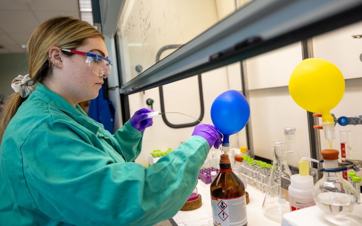 A young woman wearing a green lab coat and purple gloves dips an instrument with a long wire probe into a brown bottle with a small blue balloon attached to its mouth. 