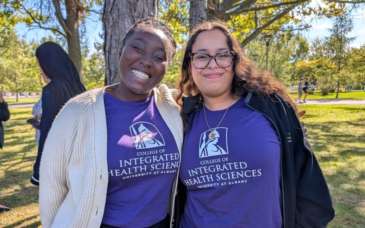 A portrait of two students wearing purple College of Integrated Health Sciences Shirts. They are outside, with trees and grass behind them.