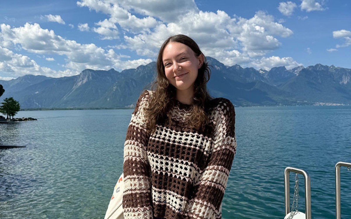 Emma standing near a body of water with a mountain range in the background somewhere in Switzerland.
