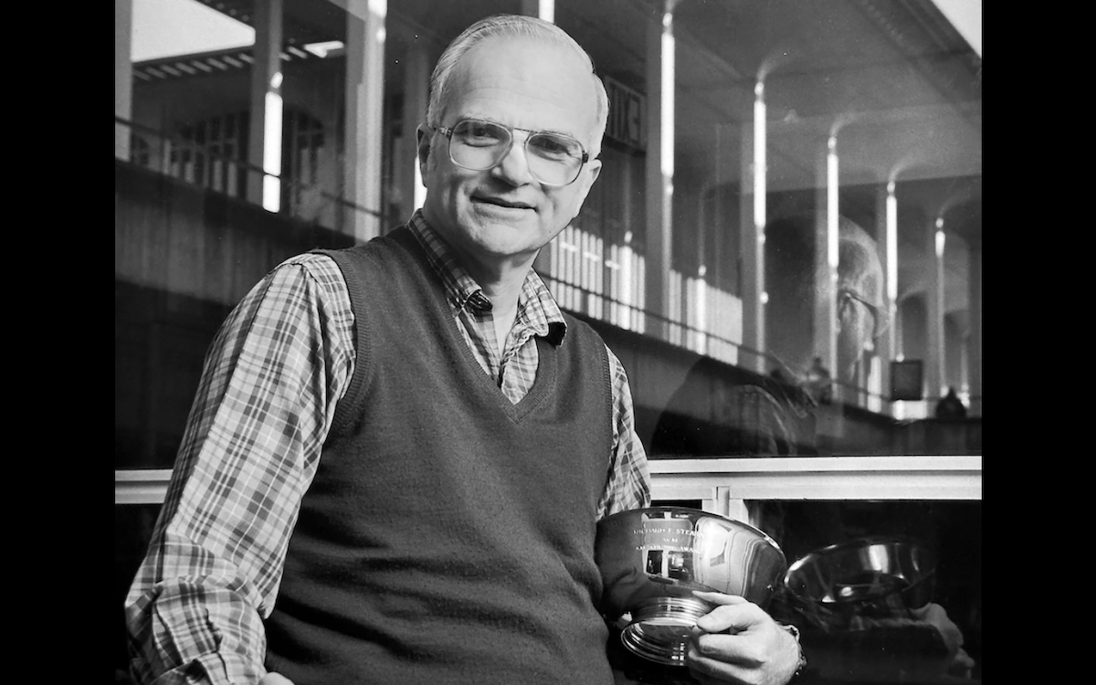 A man in a plaid shirt, sweater vest and glasses holds an award and poses for a photo