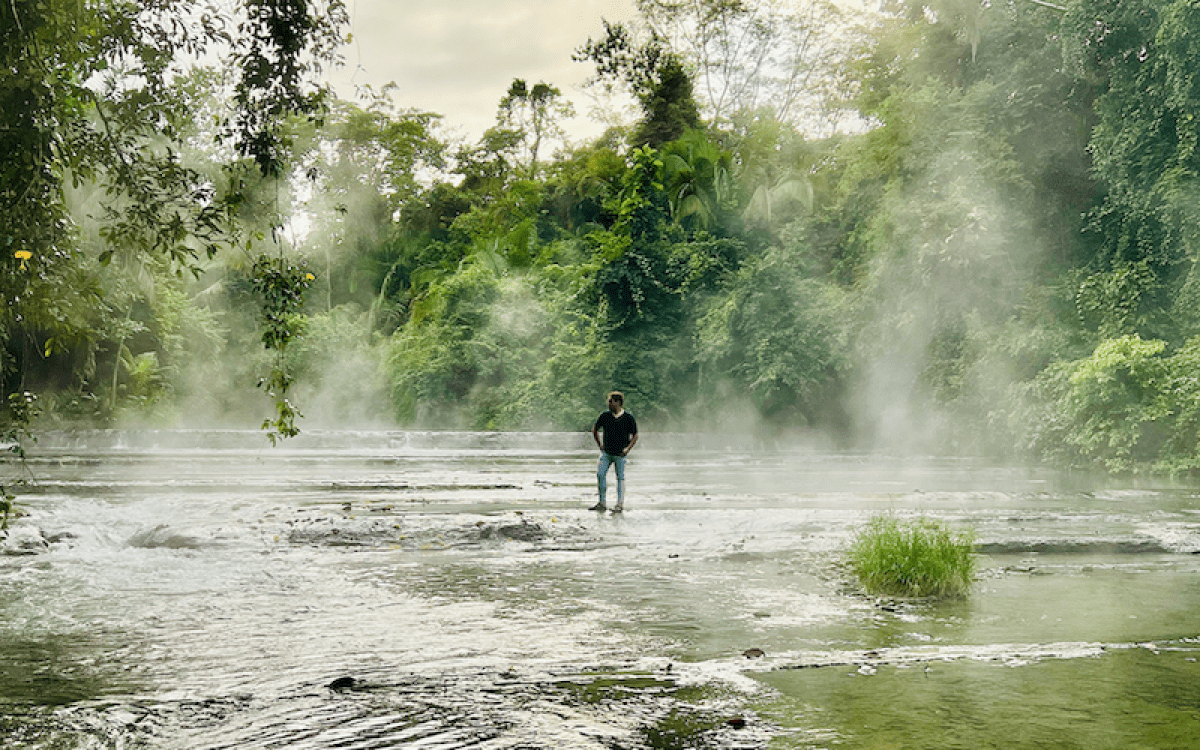 A man stands on a rock in a steaming river flowing through a jungle.