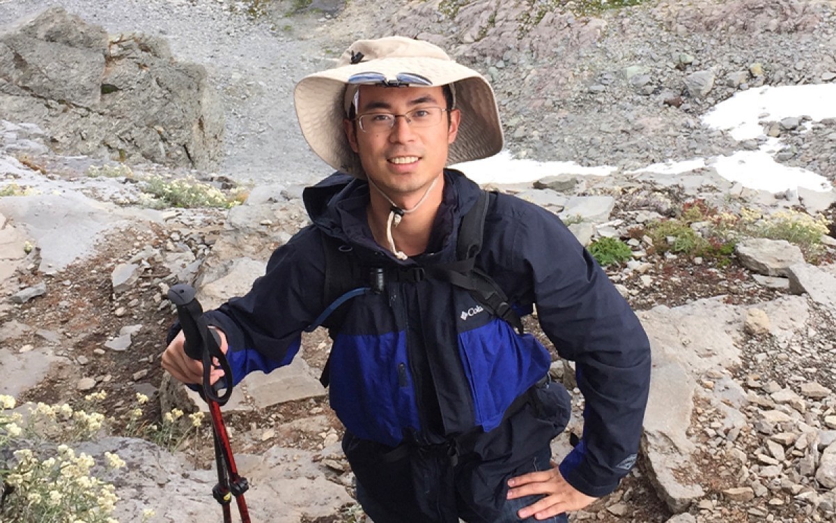 A man stands on a rock formation wearing a dark blue jacket and floppy hat, holding walking sticks.