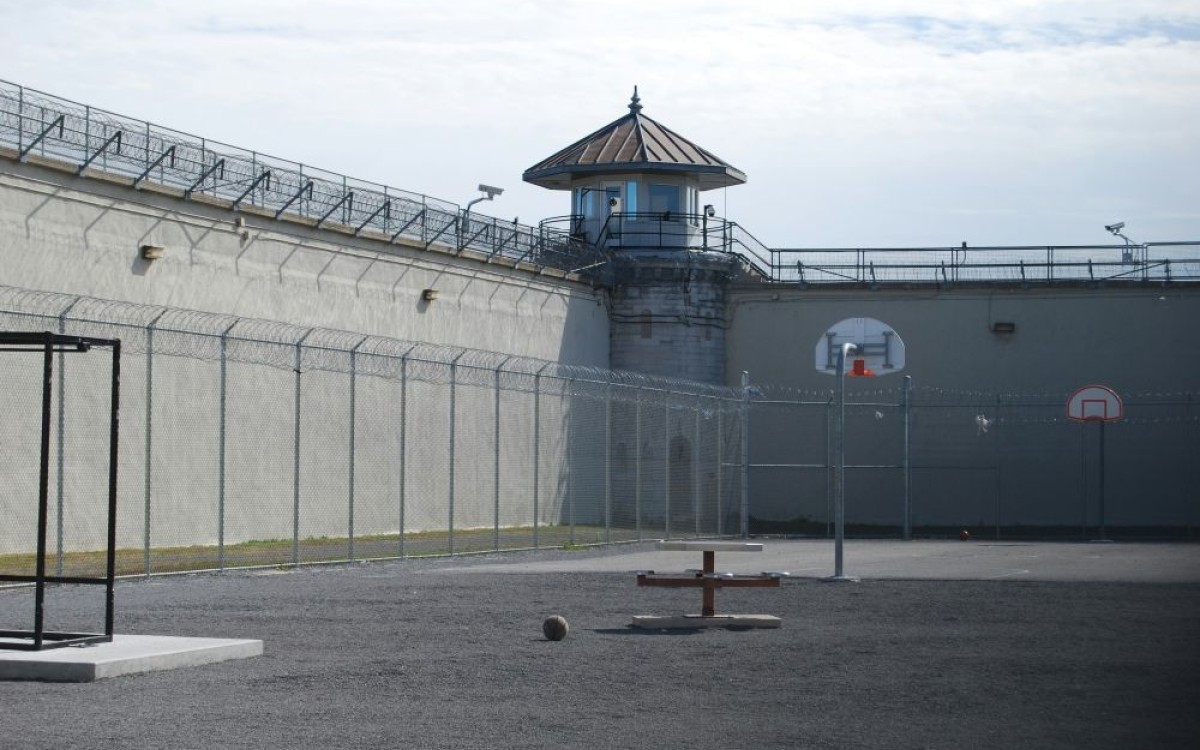 an empty prison yard surrounded by chain-link feancing and a high wall, both topped with razor wire.