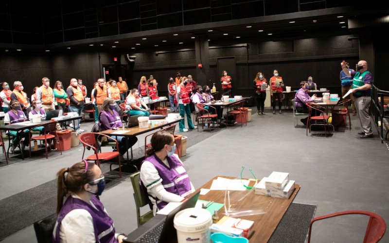 Volunteers in purple and orange vests sit at tables, listening to instructions, at the start of a vaccination clinic