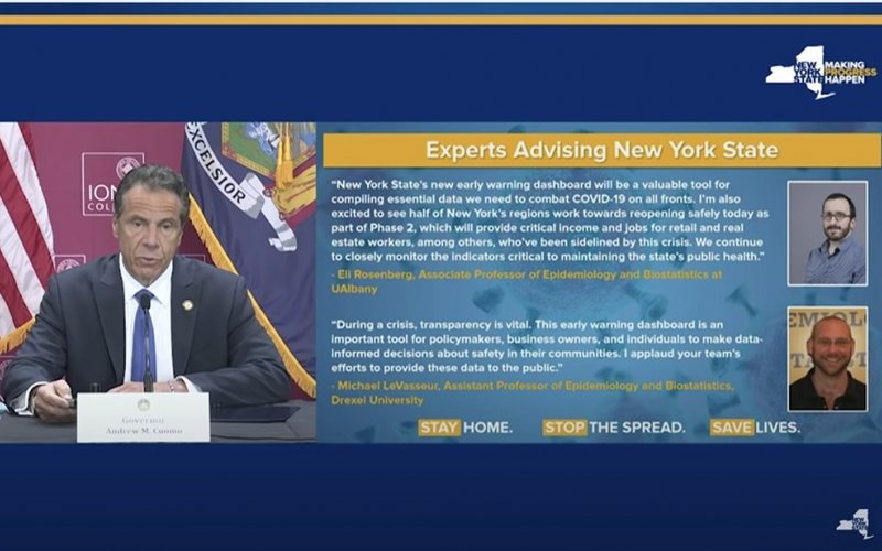 A blue screen holds a screenshot of a slide taken from Gov. Cuomo's press briefing. The slide has the heading "Experts Advising New York State" in a yellow box at the top. Below the heading is a short paragraph on Dr. Eli Rosenberg from UAlbany. Governor Cuomo is also on the left side of the screen, wearing a suit and sitting in front of New York flags.