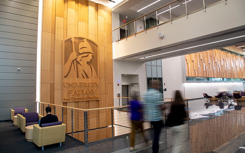 People walk through a hallway in UAlbany's ETEC building. A logo of Minerva that reads, "University at Albany/State University of New York" is displayed on the wall.
