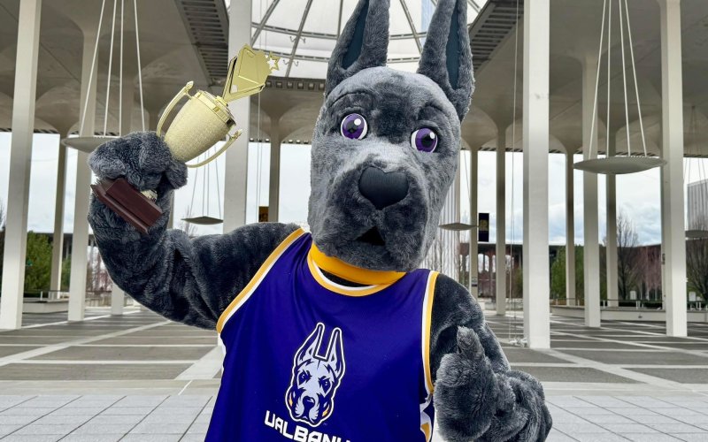 Damien the Great Dane, UAlbany's mascot, holds up a trophy while raising one finger to signify UAlbany is Number 1.