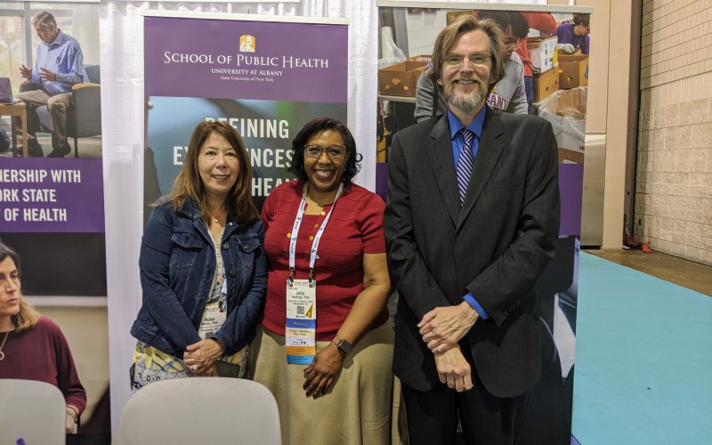 Drs. Hosler, Hastings, and Holtgrave stand behind UAlbany's table at the APHA expo, smiling.