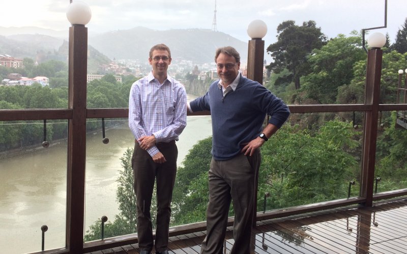 Mark Kuniholm and John Justino stand on a bridge in Georgia. Behind them is a river and mountains dotted with houses.