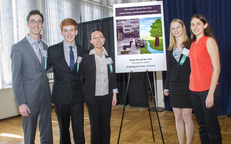 Students take photo in front of poster at UAlbany awards competition for NYS high school students in science.