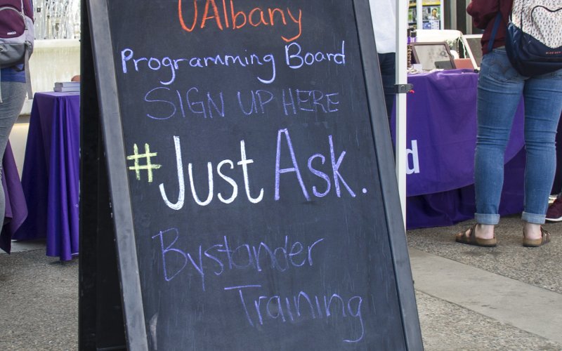 Chalkboard that says "Sign up Here, #JustAsk Bystander Training" with students in background.
