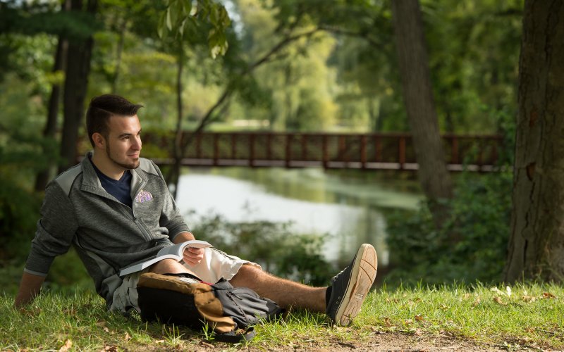 A male student sits on the grass in front of Indian Pond on campus, reading a book