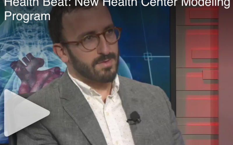 Dr. Eli Rosenberg wearing a grey suit with the health beat set behind him.